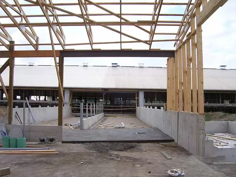 A construction site showing a steel I-beam supporting wooden roof trusses over a concrete foundation.