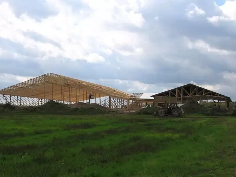 A wide view of a farm under construction featuring a large wooden truss barn frame, a tractor, and a finished metal building.
