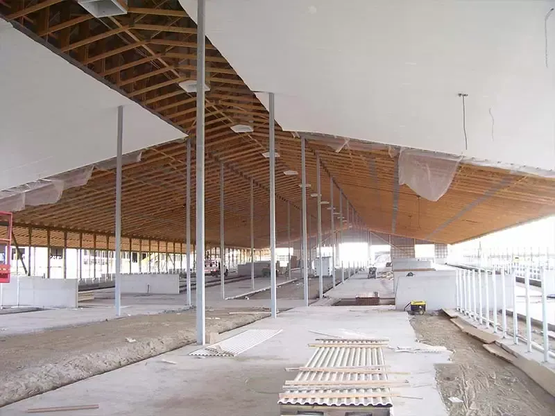 Interior of a large agricultural building showing steel support columns, wooden roof trusses, and white ceiling insulation panels.