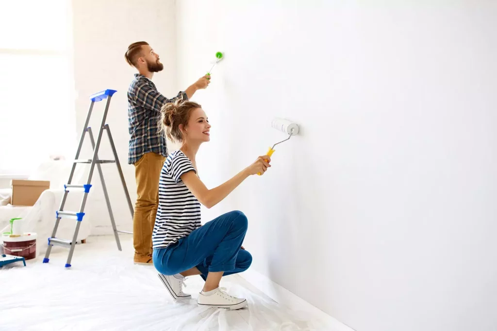 A man and woman smiling while painting a bright white interior wall with rollers in a room prepared with drop cloths and a ladder.