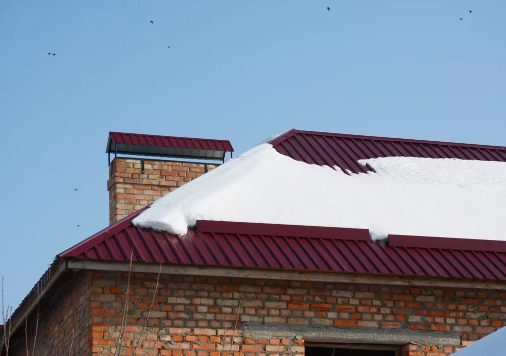 A brick house featuring a deep red corrugated metal roof with a significant snow load and a matching chimney cap against a clear blue sky.