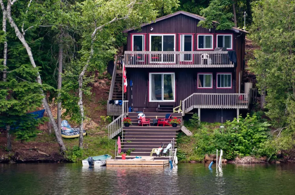 A two-story dark wood lakeside cottage with multi-level decks, a dock, and a boat on a forested shoreline in the Great Lakes region.