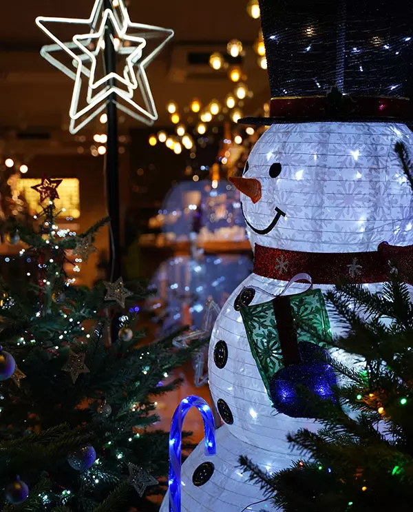A brightly lit white snowman decoration with a top hat and scarf, surrounded by glowing Christmas trees and neon stars at night.