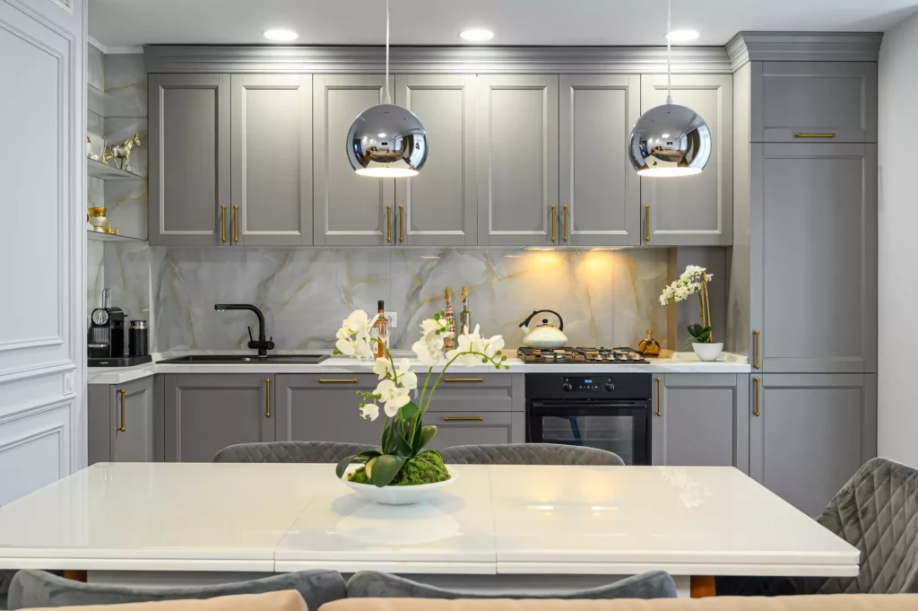 A modern kitchen featuring sleek grey shaker-style cabinets with gold hardware, a marble backsplash, and a clean white dining table.