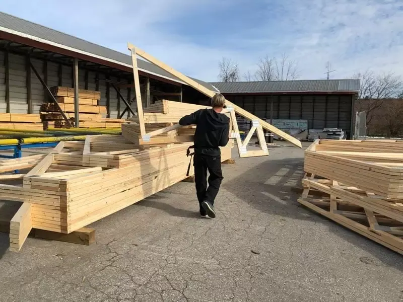 A carpenter at Great Lakes Rona carrying a wooden roof truss in a lumber yard.
